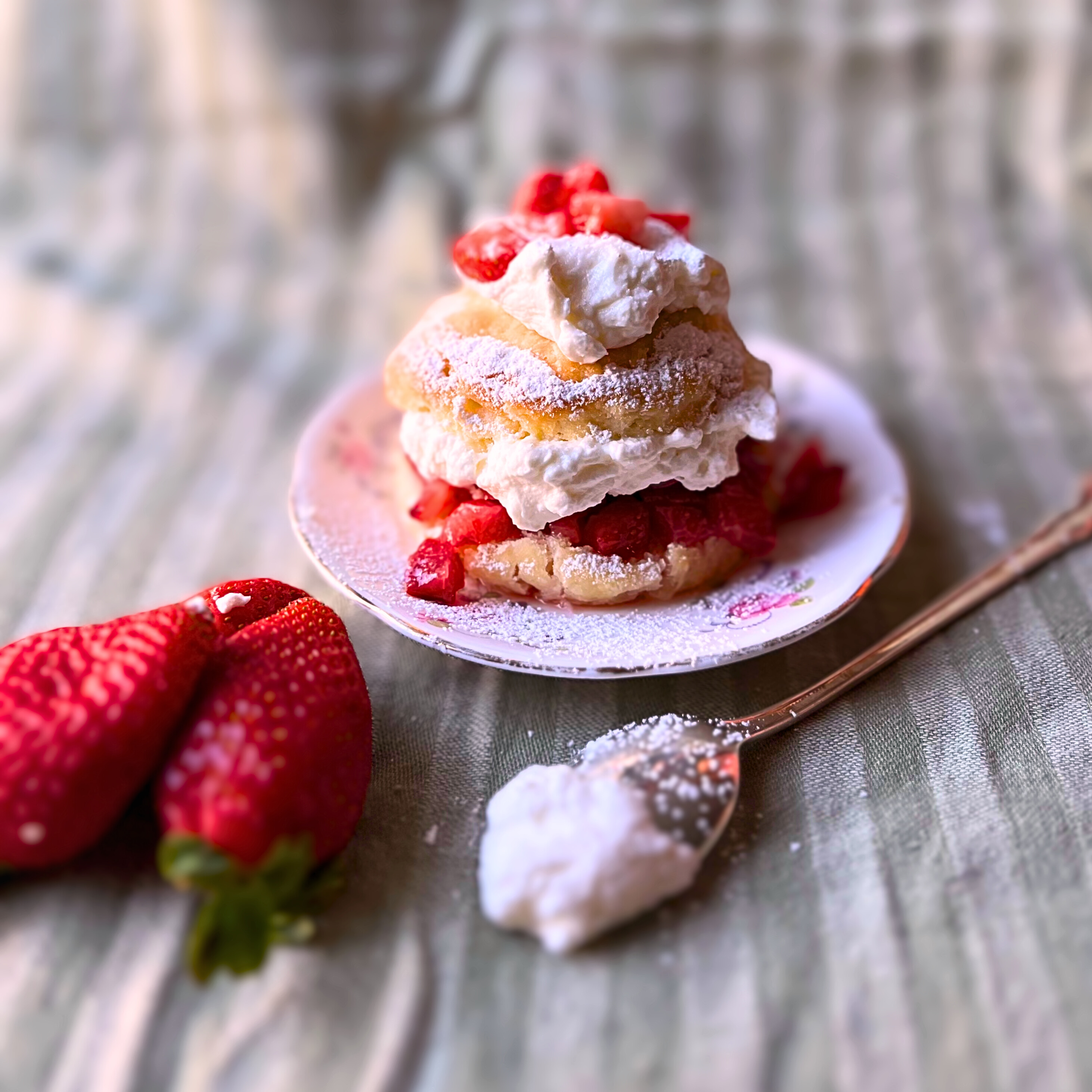 Rich and flaky butter milk biscuits with sweetened strawberries and whipping cream. on a pink floral plate. Whole strawberries and a spoon with whipping cream in front.
