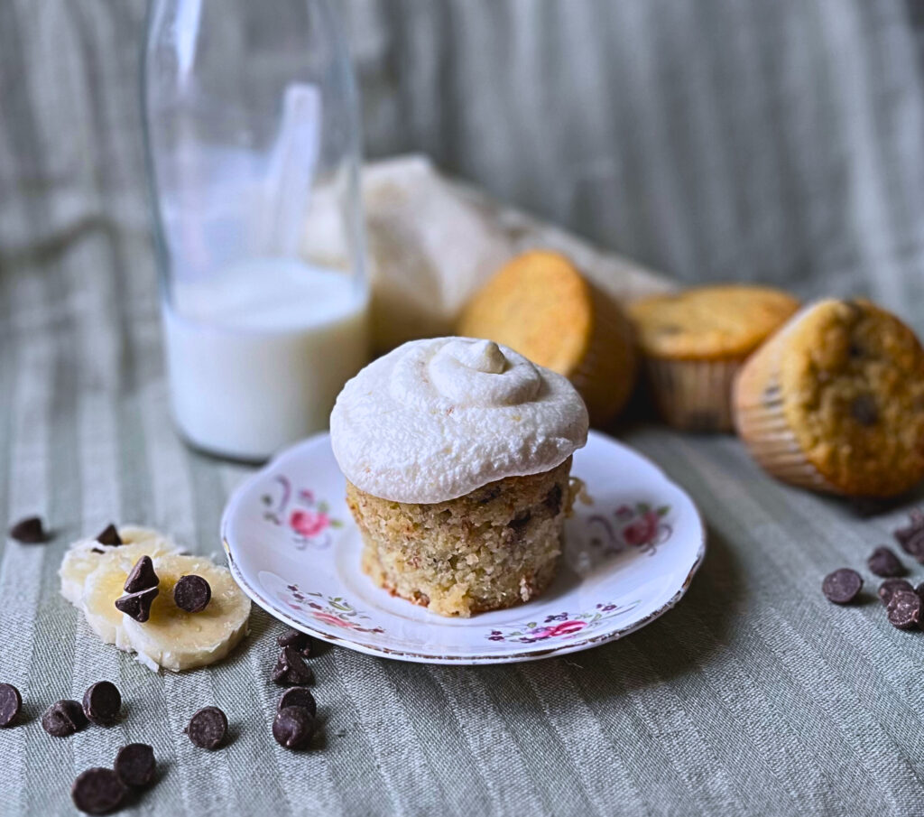 Wholesome banana chocolate chip cupcake with Banana icing on top on a floral plate with three cupcakes, milk and an icing bag in the background. Beside is sliced banana and chocolate chips.