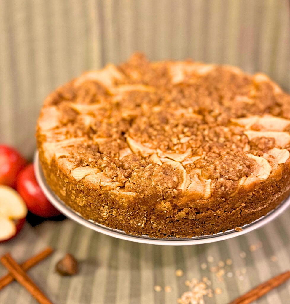 Apple crisp cheesecake on a cake stand with apples cinnamon sticks and an acorn