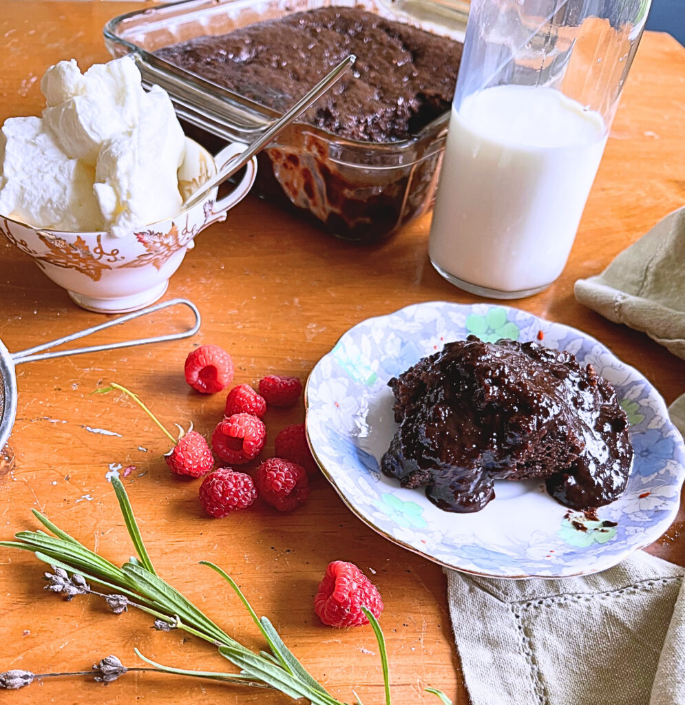 Elizabeth's Fudgy chocolate pudding cake on a blue floral plate. In the background the rest of the cake a bottle of milk and a cup of cream. In the foreground, raspberries and lavender.