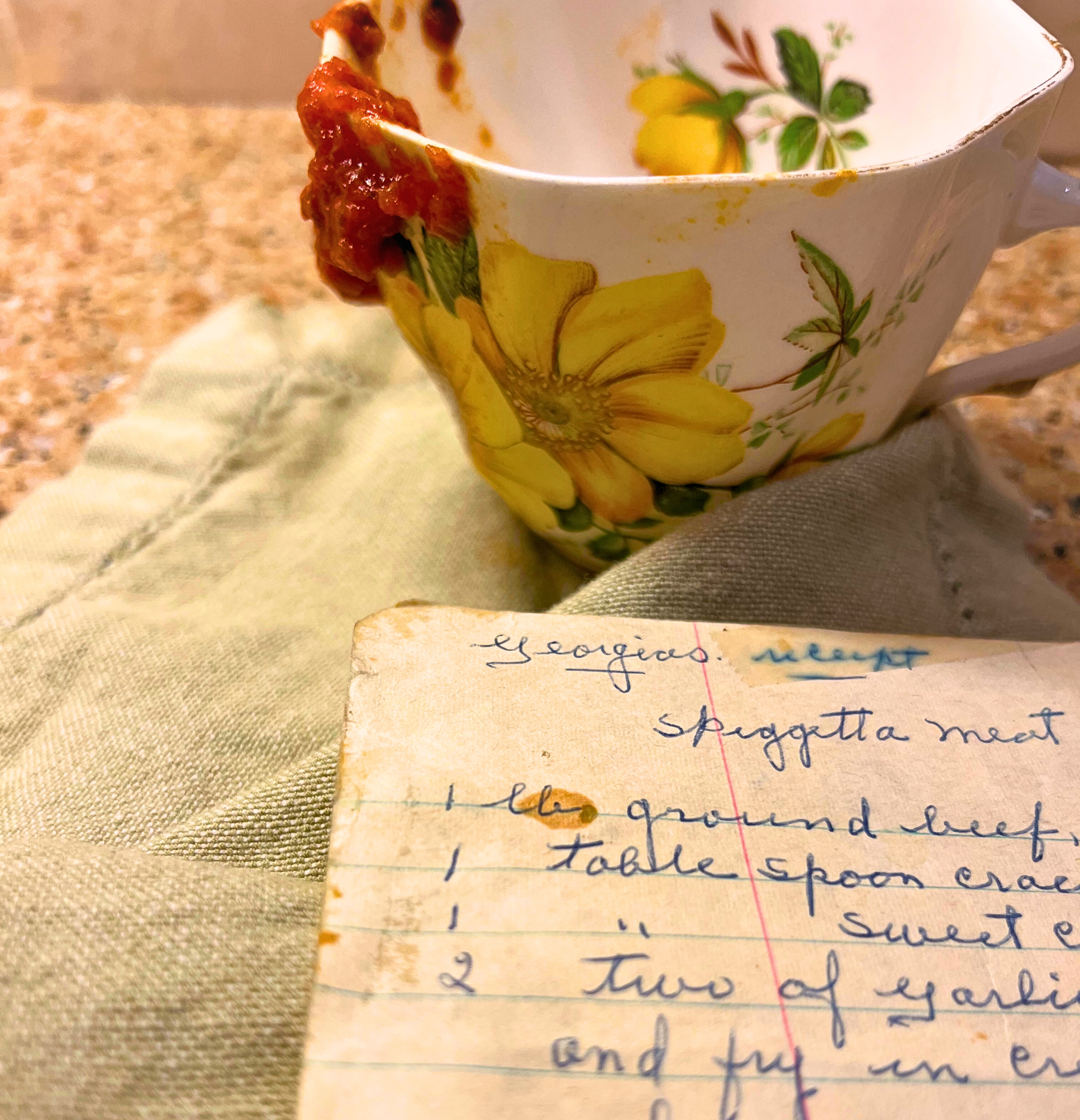 A yellow floral teacup with marinara sauce running down the side on a green/grey napkin. in front is a hand written recipe.