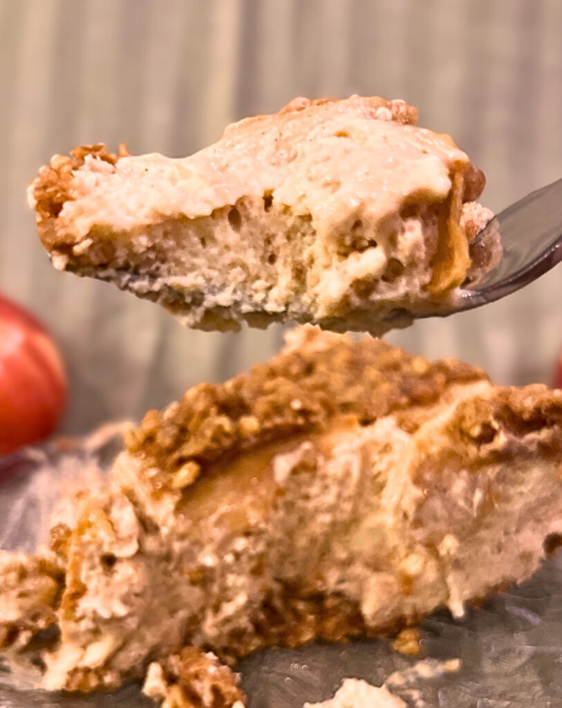 A fork holding up some Apple crisp cheesecake with a slice of the cake on a glass plate in the background