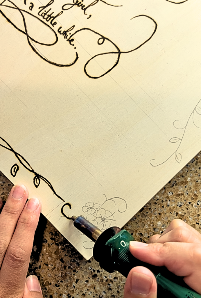 A woman holding a wood burning tool and using it on a wooden canvas to trace flowers and vines.