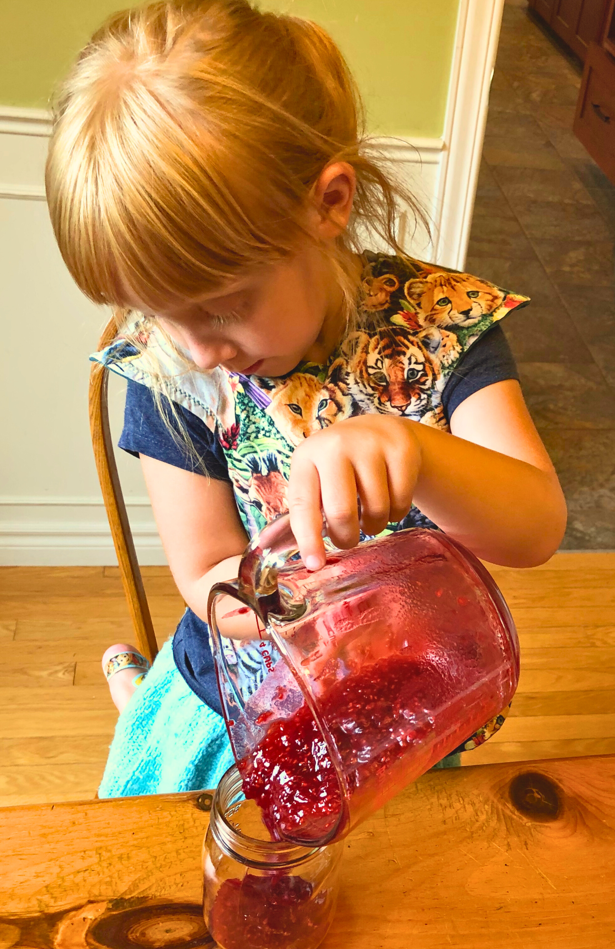 A little girl pouring raspberry jam into a jam jar.