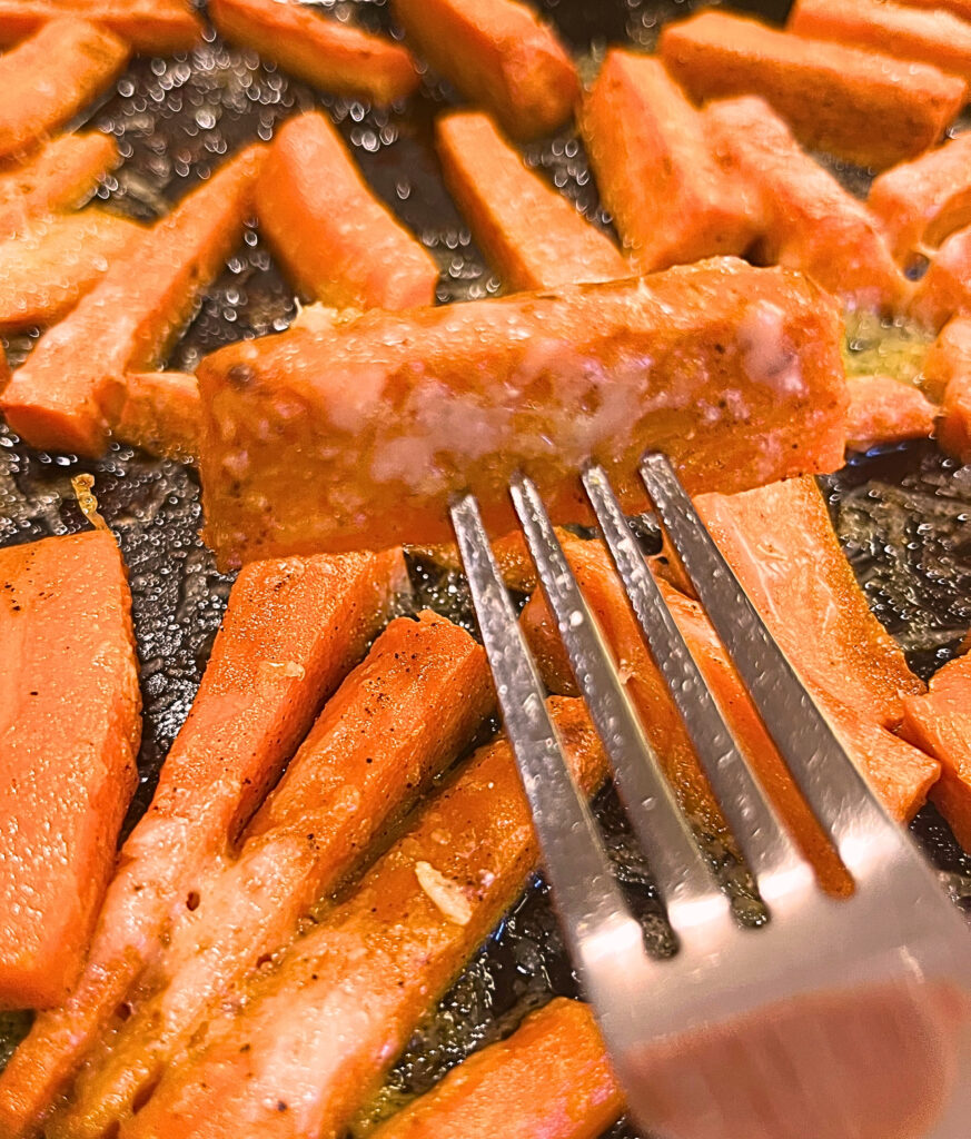 A metal fork holding up a Oven sauteed carrot with cheese and a tray of carrots in the background.