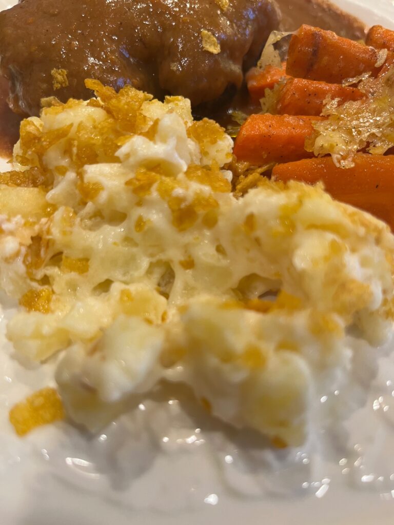 Cheesy hashbrown casserole on a white plate with Salisbury steak and cooked carrots in the background.