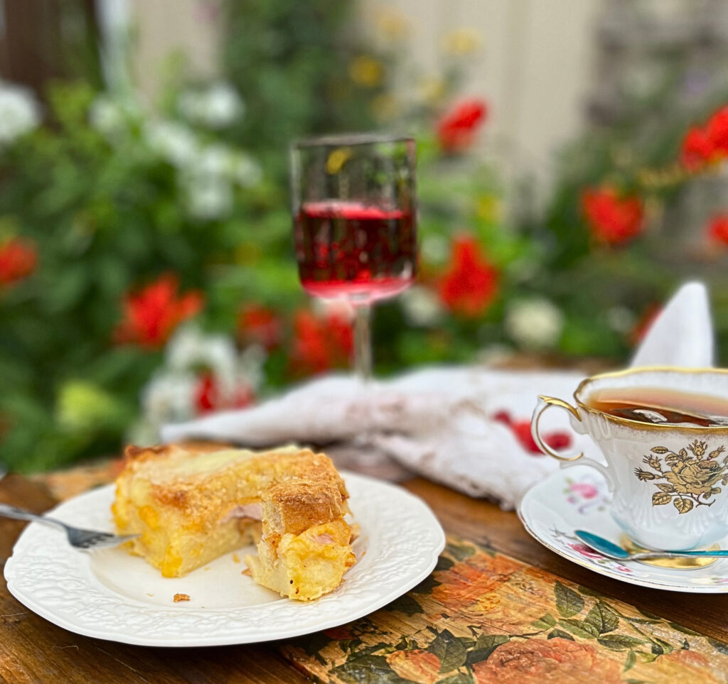 An outdoor table with Make ahead breakfast strata on a white plate, a cut of tea and some cranberry juice in the background.