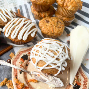 A muffin with white icing on a brown floral plate. There is a piping back with white icing beside and more muffins in the background.