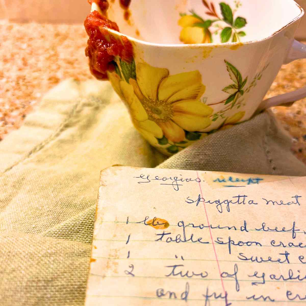 There is a yellow floral cup with pasta sauce on it. In front is a recipe card.