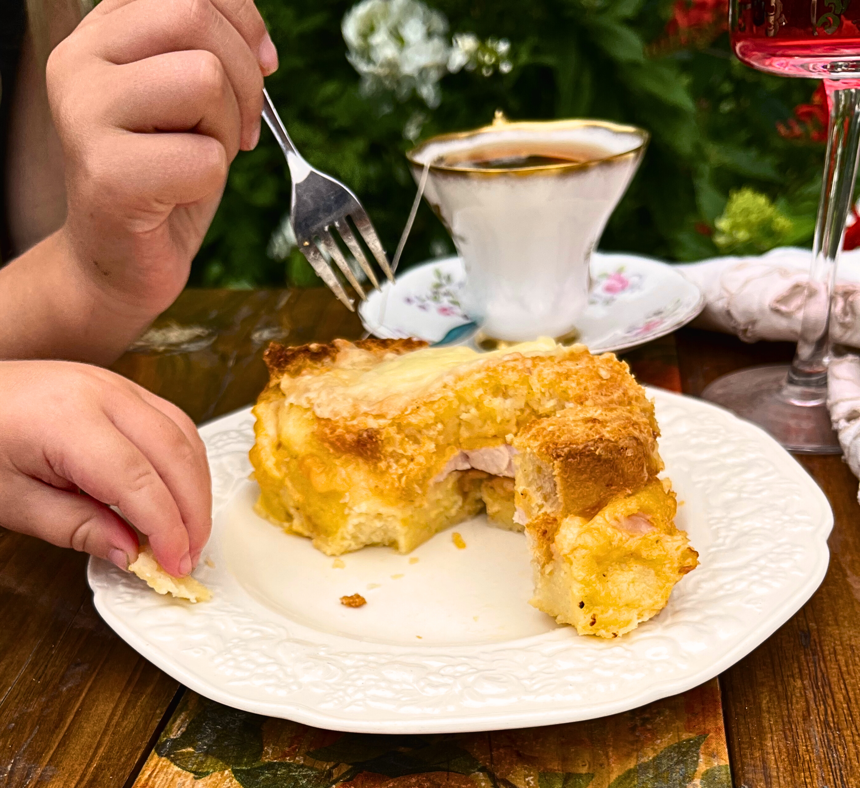 Outdoor table with make ahead breakfast strata on a white plate with a cup of tea in the background and two children's hands eating it.