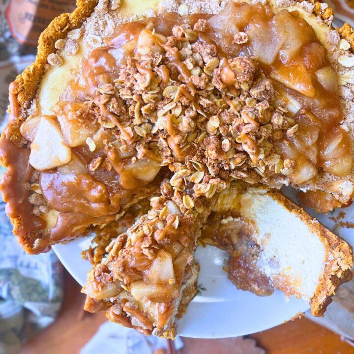 A baked apple crisp cheesecake with a cooked apple mixture and a crumbly oatmeal crisp layer on top. the cake is sliced. Once slice is on it's side.