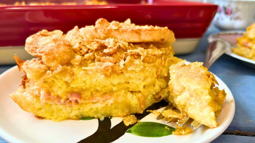 A bread, ham, and egg casserole on a plate. There is more in a casserole dish behind.