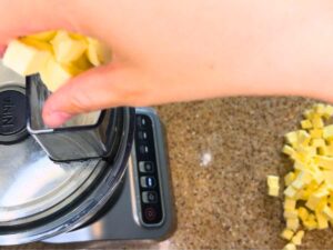 A woman is dropping cubes of butter through the funnel of a food processor.