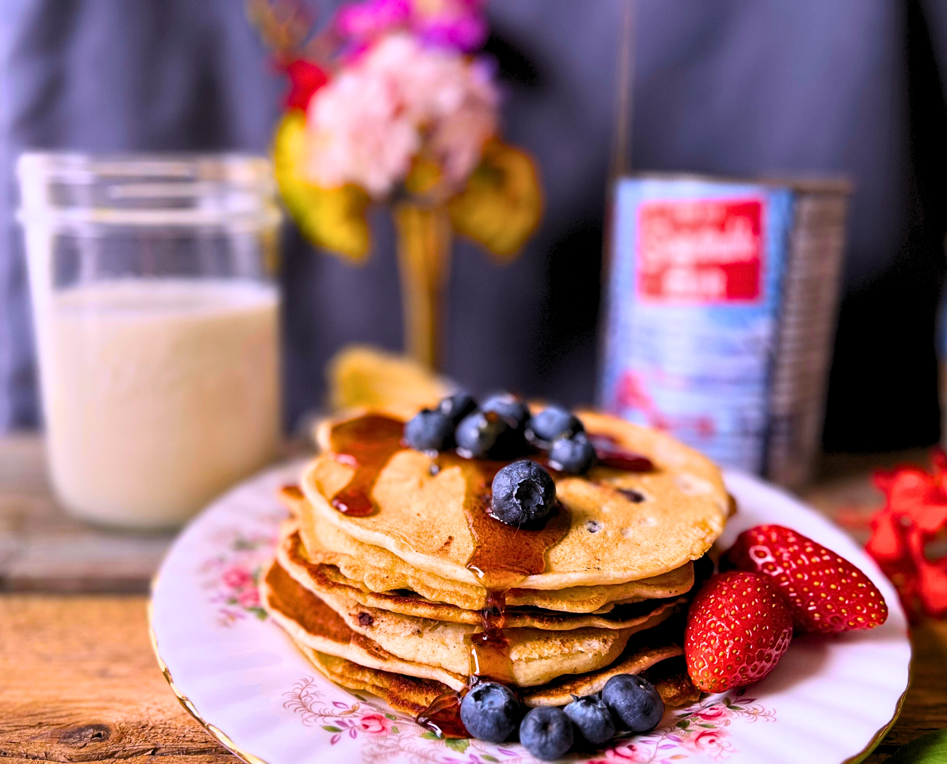 The Best Soft and Thin Pancakes with blueberries, strawberries, and maple syrup. In the background a jar of milk, a tin of maple syrup and an arrangment of flowers in a gold vase.