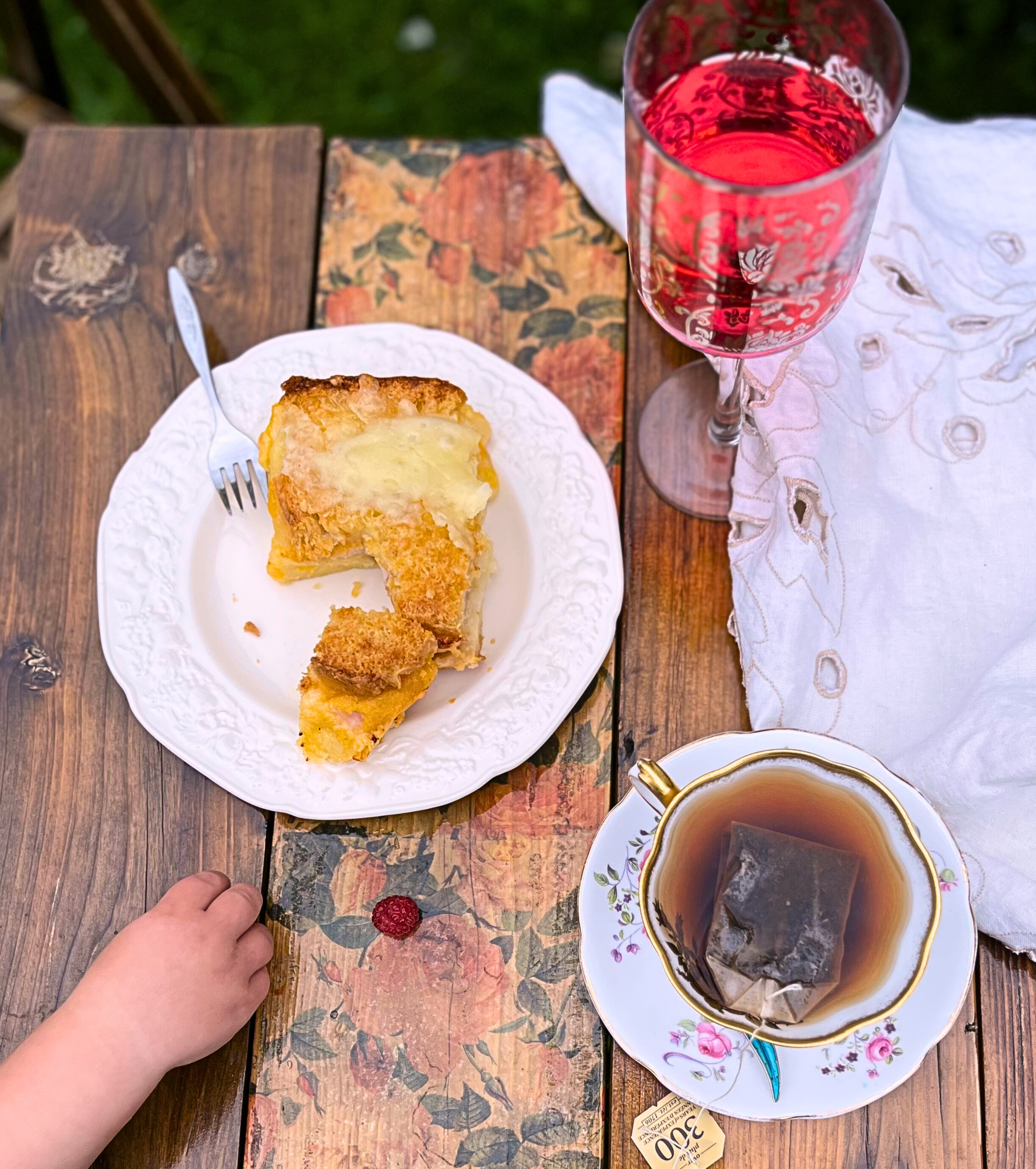 An overhead view of a wooden table with make ahead breakfast strata on a white plate with a metal fork, a cup of tea, and cranberry juice and a child's hand taking raspberries.