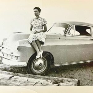 An antique picture of a woman sitting on the hood of a car.