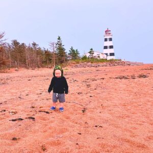 Toddler walking on the beach by a white and black lighthouse.