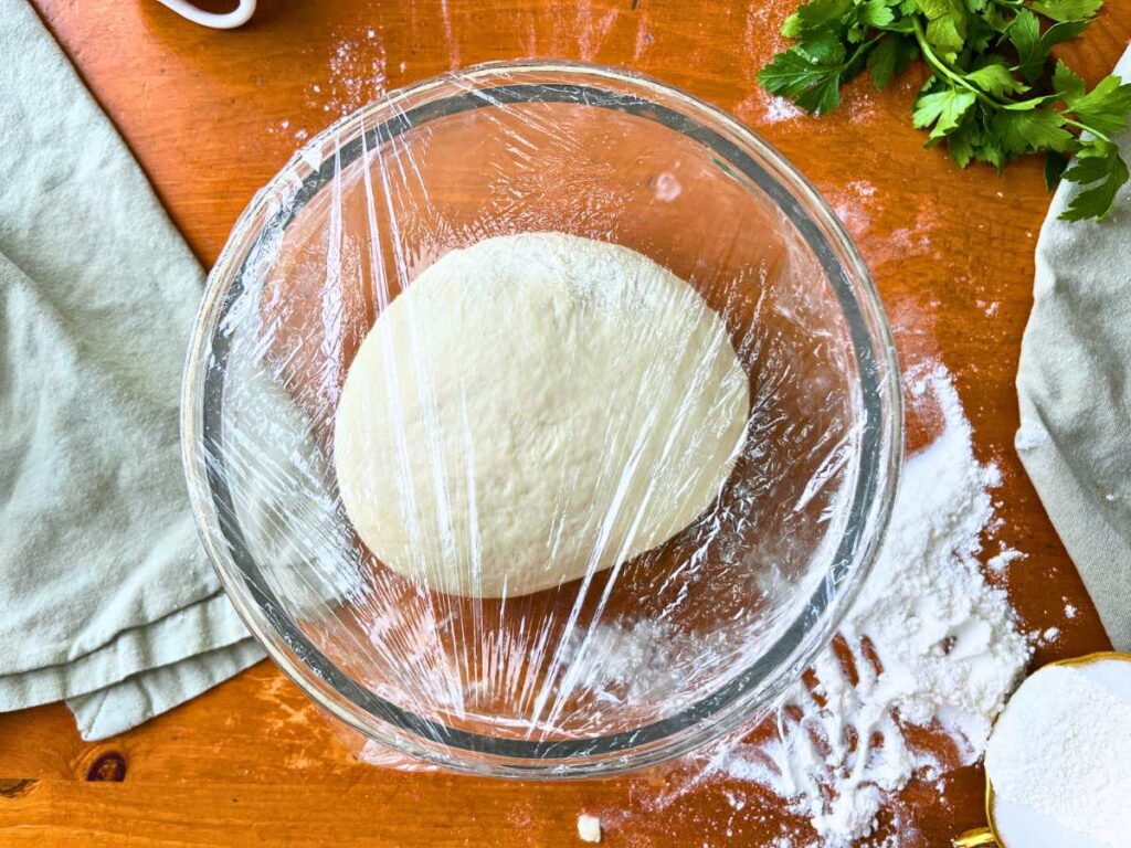 A glass bowl with pizza dough inside. It is covered with plastic wrap.