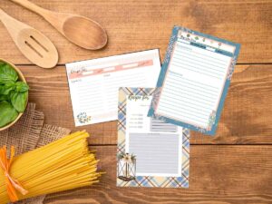 Three blank recipe cards sitting on a wooden table with dried spaghetti and basil on it.