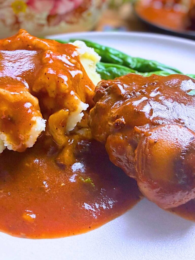A side view of a salisbury steak with mashed potatoes and gravy. There are green bean in the background.