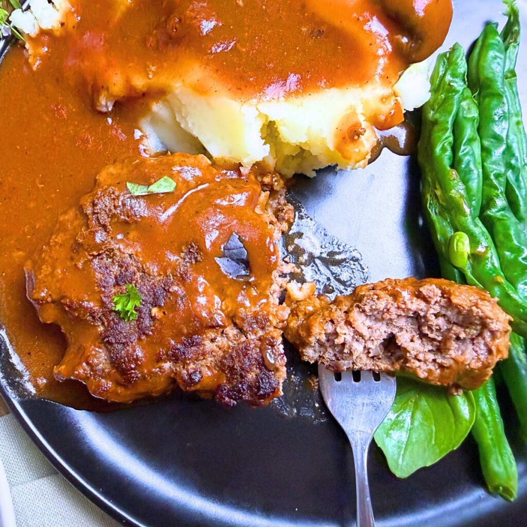 A black plate with salisbury steak, mashed potatoes, and green beans. There is a fork with a slice of salisbury steak on it.