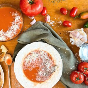 There are two bowl of tomato soup on a wooden table. Some fresh tomatoes, peppers, and garlic are around. There is grated parmesan cheese on top.