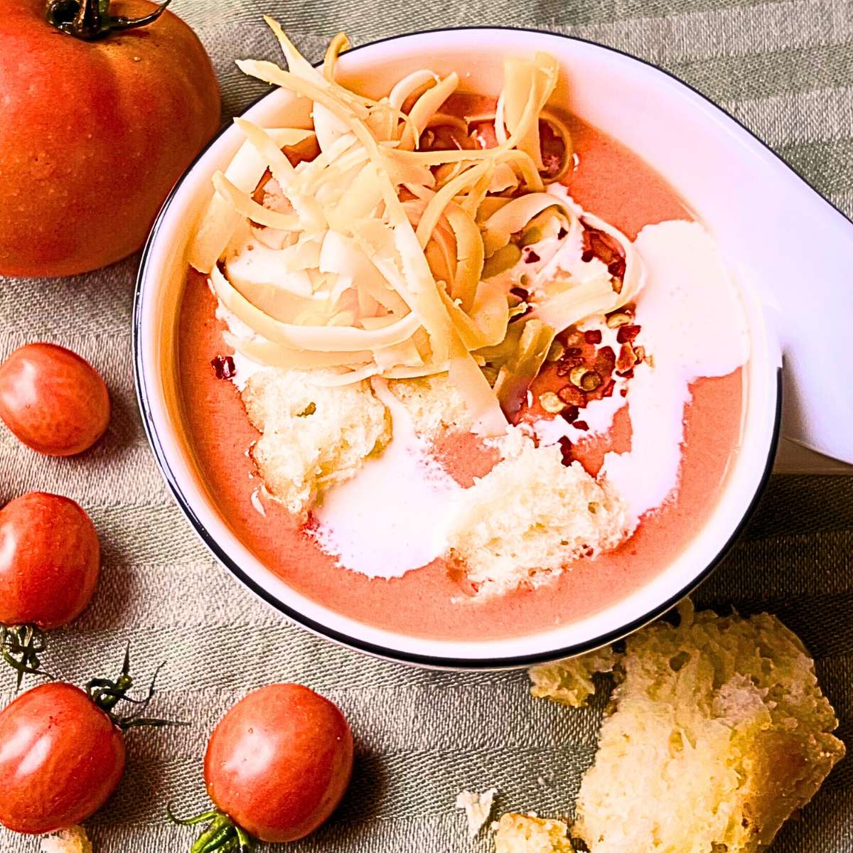 A mug with tomato soup. There is some cream, cheese and pepper flakes on top. There are tomatoes around.