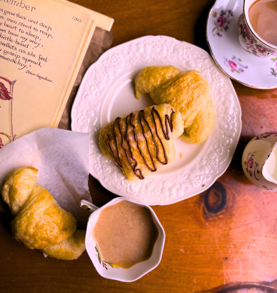 croissants and pain au chocolat on a white plate. Tow cups of coffee and an open book on a wooden table