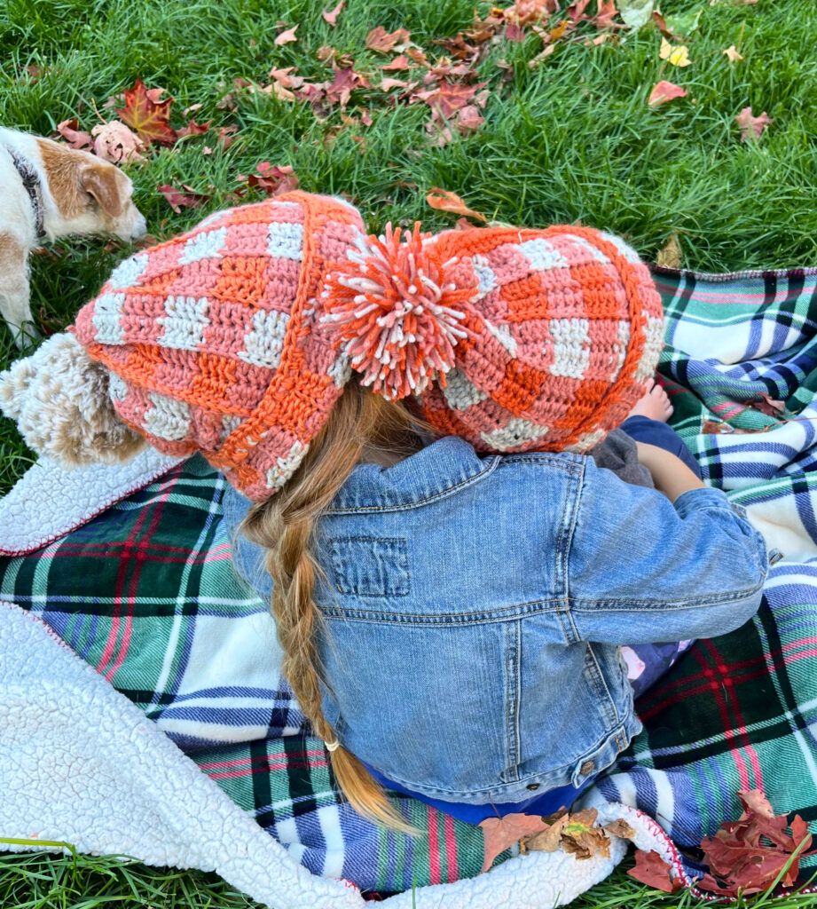 Two children and a dog sitting on a plaid blanket wearing orange crochet hats