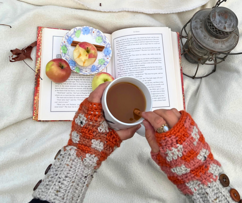 Woman wearing orange and gray fingerless gloves stirring cider with a cinnamon stick, reading a book with three apples on it, a plate and two cinnamon sticks. All of a white blanket with a metal lantern on it.