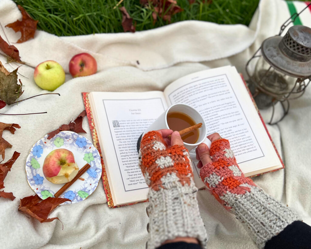 Woman wearing orange and grey buffalo plaid fingerless gloves, holding a mug of cider and a cinnamon stick. There is a book, three apples a plate and a metal lantern, all on a white Hudson's Bay blanket.