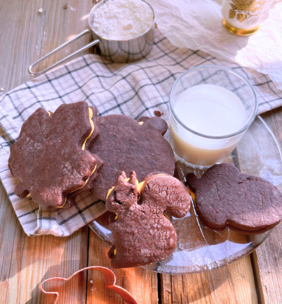 Chocolate sandwich cookies sitting on  a glass plate. A glass of milk and a grey and white kitchen towel on a wooden table with an acorn cookie cutter in the foreground.