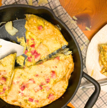 A frittata sliced in a cast iron skillet. With a small metal serving spatula and a piece of frittata on a white plate.