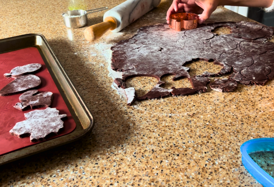 Woman using cookie cutters to cut shapes out of chocolate cookie dough. A rolling pin and measuring cup of flour in the background. A lined tray with cut out cookie shapes off to the side.