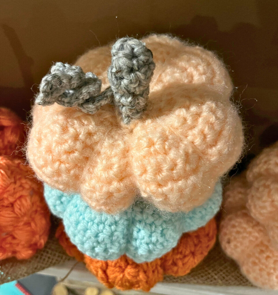 A stack of crochet pumpkins in orange, blue, and peach. Surrounded by more pumpkins on a piece of burlap