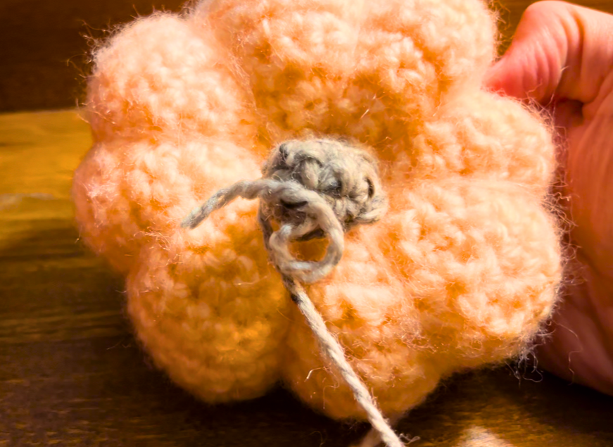 A peach yarn crochet pumpkin from above with gray yarn attatched