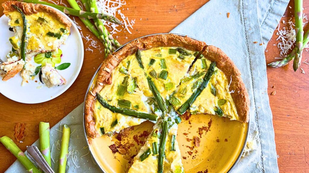 A quiche that is sliced into portions. There is asparagus and grated cheese of to the side.