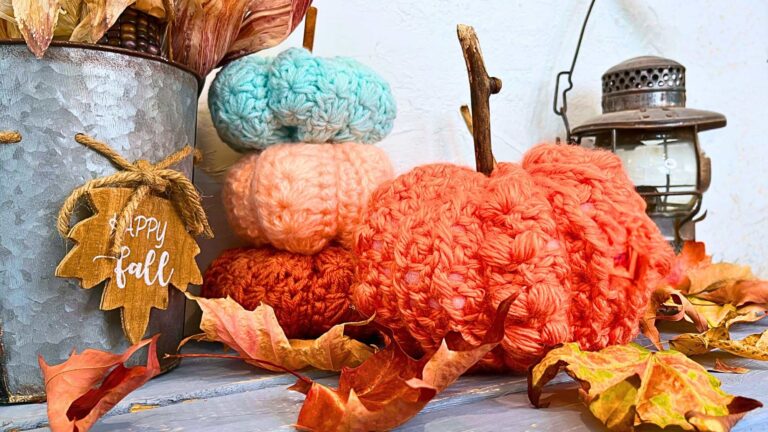 Crochet pumpkins on a blue table there is a metal basket with corn inside and leaves around.