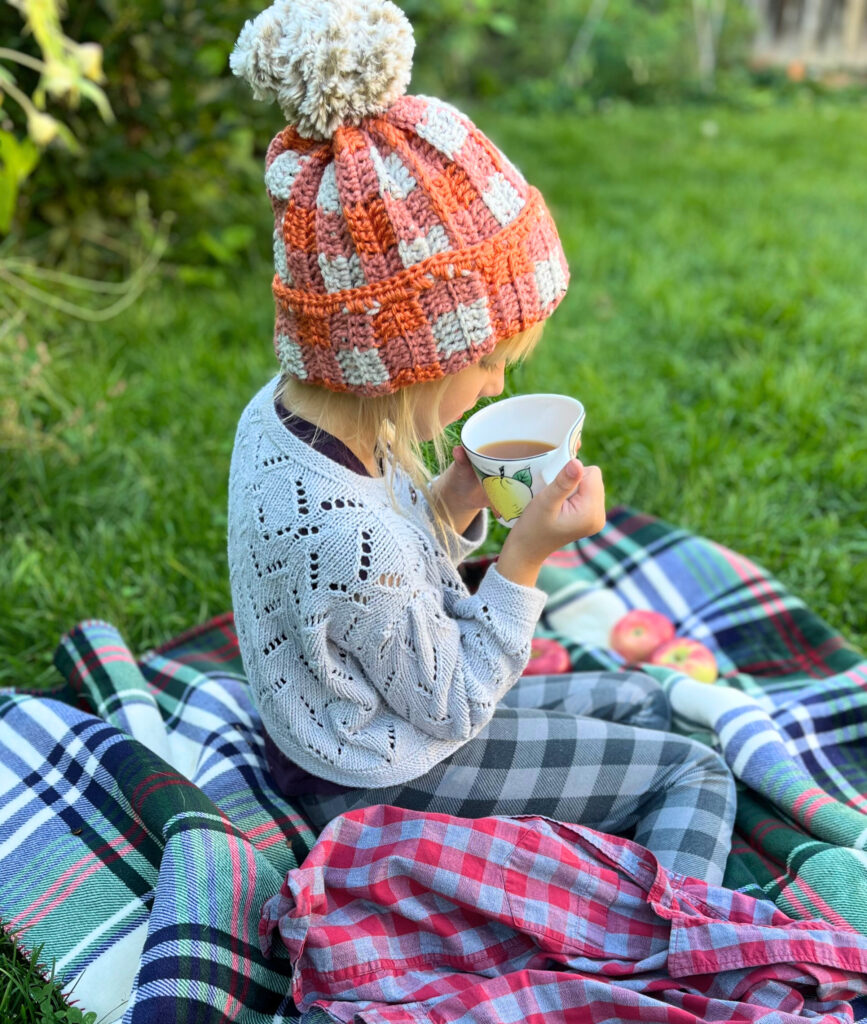 girl with white and orange crochet hat sitting on a plaid blanket, drinking cider.