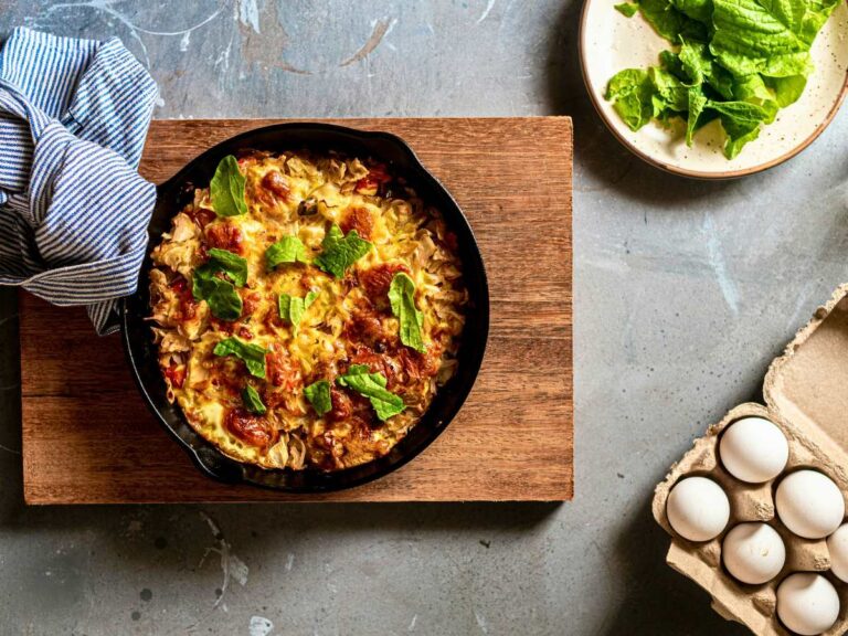 A skillet of frittata on a wooden cutting board. There is a carton of eggs and a plate of lettuce.