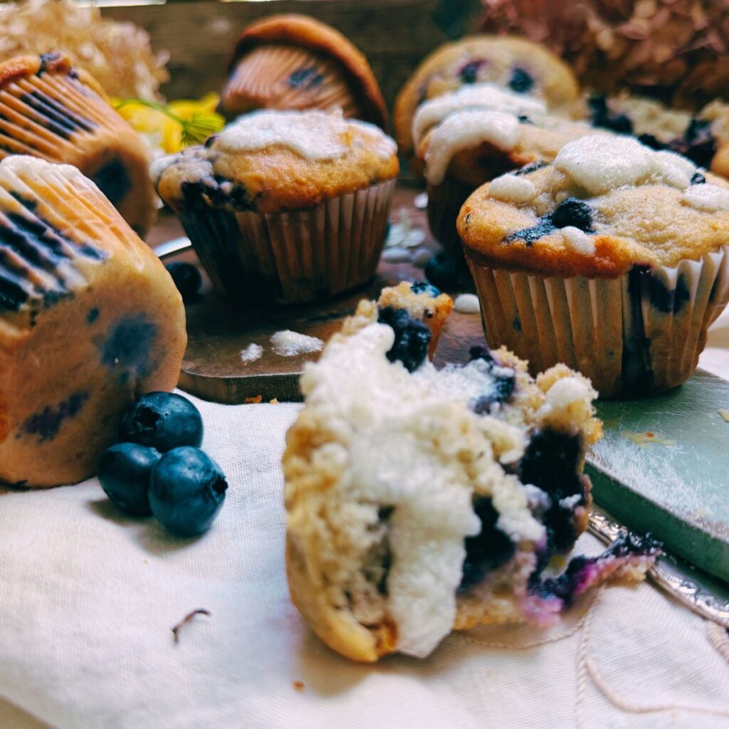 A collection of blueberry muffins. Some have a vanilla glaze. One is split in half.