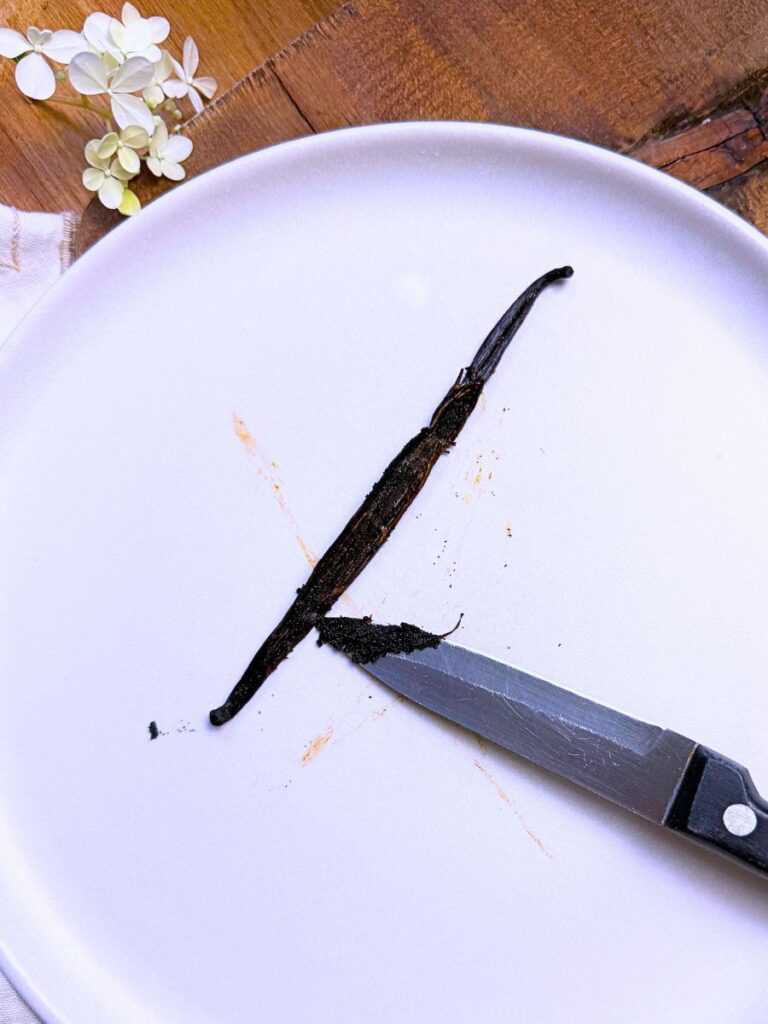 A vanilla bean on a white plate. It has been split with a knife and seeds removed.