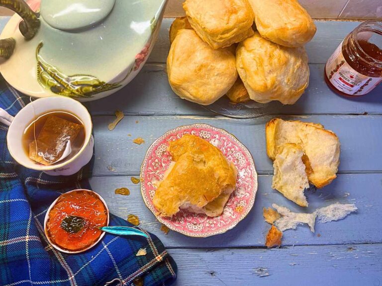 There is a Scottish butter roll on a pink plate. There are more in the background. There is jam beside and a cup of tea and teapot.