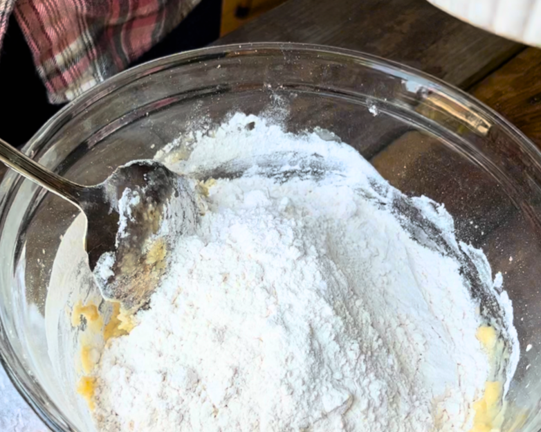 Woman mixing more flour into bread dough, in a glass bowl with a metal spoon