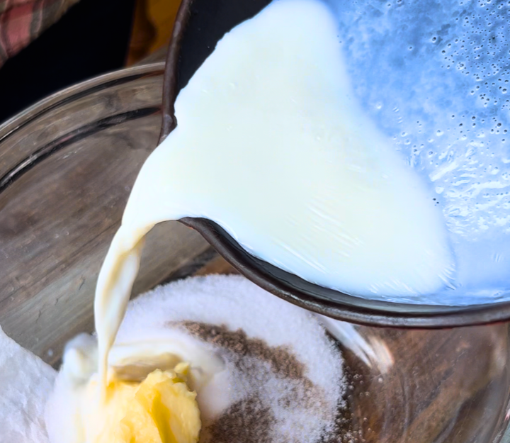 Pouring hot milk from a pan into a glass bowl with sugar, salt, butter, and cardamom.