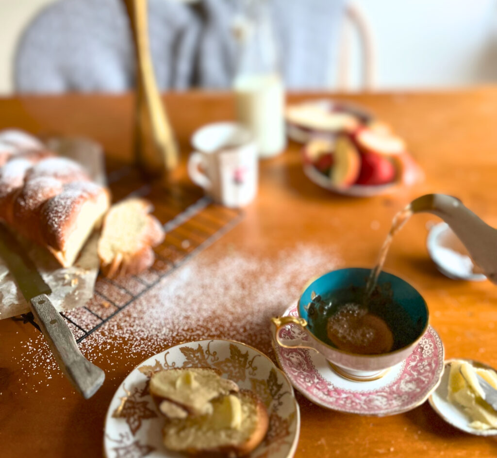 Tea set up on a wooden table, with a pulla bread loaf on a wire rack, slices of pulla on two plates, two cups of tea, a plate of strawberries and apples, a small bowl with butter and a knife, a jar of milk, a vase, and a teapot pouring tea.