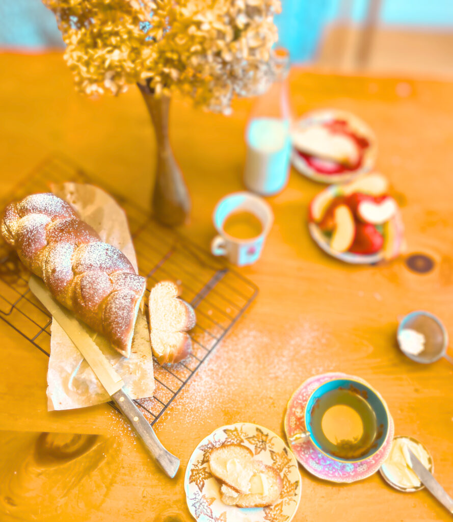A cut loaf of Pulla bread on a wire rack with two cups of tea, two plates with slices of bread on it, a jar of milk, plate of apples and berries, small plate of butter and a knife and a vase of dried hydrangeas.
