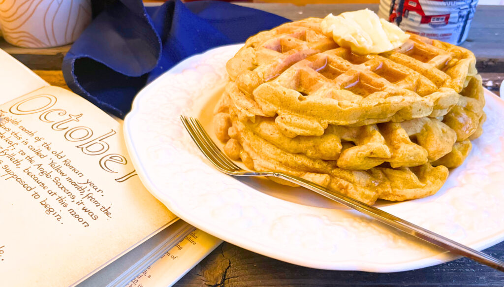 A plate of pumpkin waffles with a fork, butter on top, and maple syrup. There is an open book beside and a blue napkin in the background.