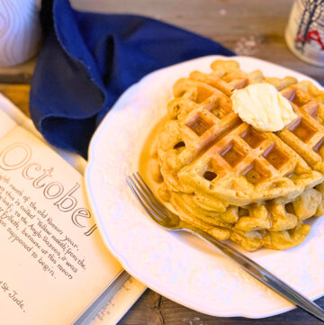 A stack of pumpkin waffles on a white plate with butter and maple syrup. The plate is sitting on a blue napkin with a mug in the background and an open book beside.
