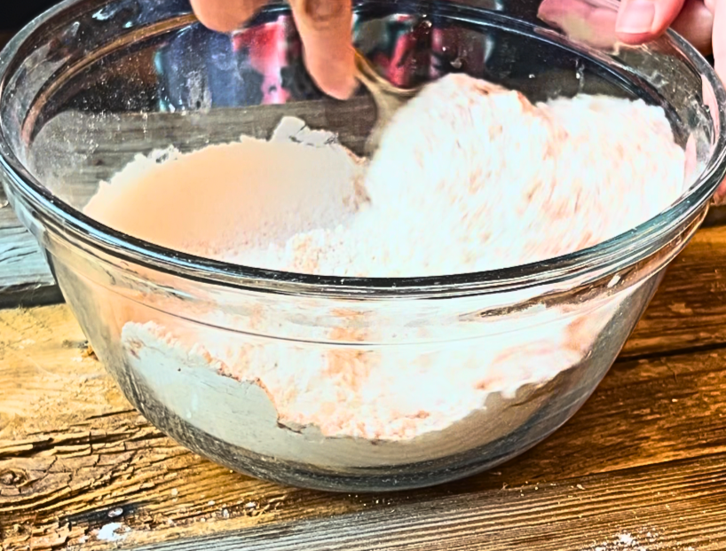 A glass bowl with flour, baking powder, salt, and cinnamon being mixed with a metal spoon.